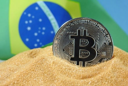 Moeda de bitcoin na areia com a bandeira do Brasil no fundo