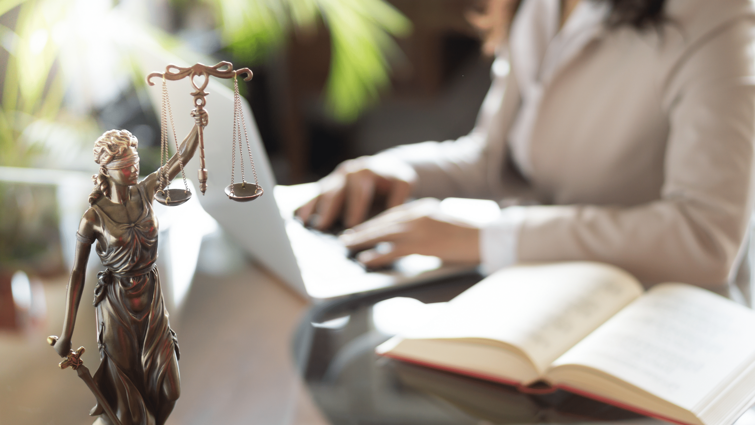 mulher sentada com um livro de direito penal e digitando em um computador. Simbolo da justiça a frente de imagem.