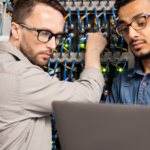 Dois administradores de sistemas trabalhando em um servidor, representando o artigo sobre as '5 razões para usar Linux em servidores ao invés de Windows Server'. A imagem mostra profissionais em um ambiente técnico, com cabos e equipamentos de rede ao fundo.
