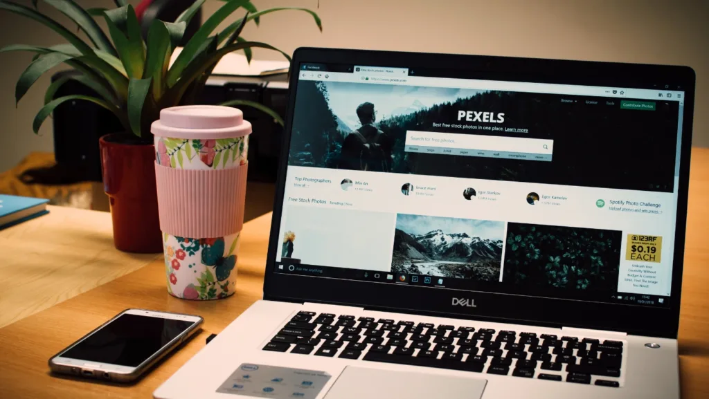 Notebook Dell com site de imagens Pexels aberto, copo decorado e planta sobre mesa de madeira