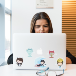 Pessoa usando um notebook adesivado com logos do GitHub, trabalhando em um ambiente de escritório com grandes janelas ao fundo.