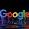 Logotipo 3D do Google em cores vibrantes sobre uma superfície reflexiva, com fundo preto.