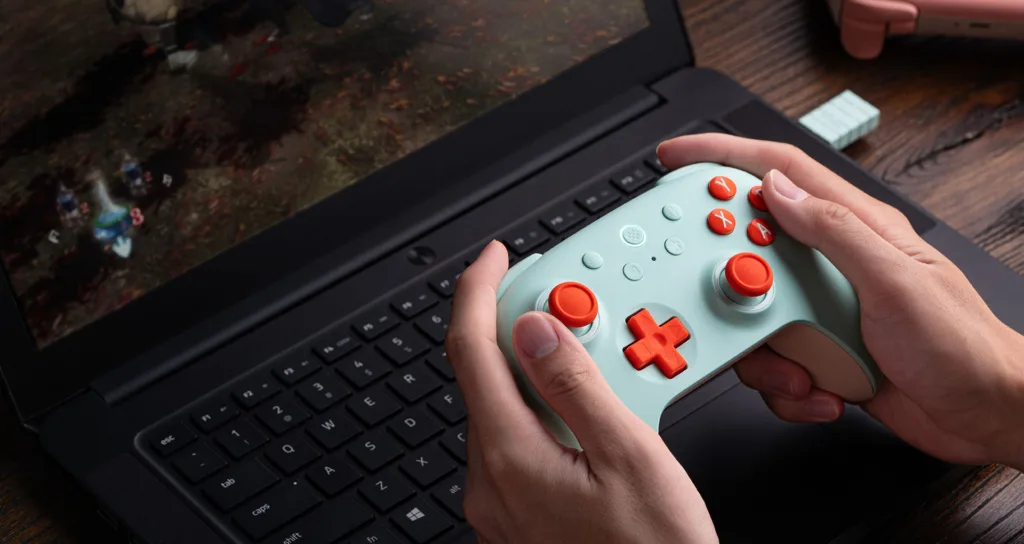 Pessoa segurando um controle sem fio 8BitDo Ultimate 2C em cores verde e laranja, jogando em um laptop com o jogo em exibição na tela.