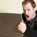 Homem expressando raiva enquanto segura um notebook, parecendo gritar ou reclamar em frente à tela.
