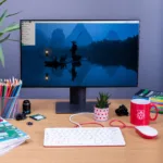Mesa de trabalho com monitor mostrando a interface do Raspberry Pi OS, teclado e mouse com o logotipo do Raspberry Pi. Há canetas, uma xícara de café e livros ao redor.