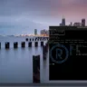 Captura de tela do desktop do Redox OS exibindo um terminal aberto. O terminal mostra o login do sistema com o usuário user e uma interface gráfica minimalista com uma barra de aplicativos na parte inferior da tela. A imagem de fundo apresenta uma paisagem urbana à noite.