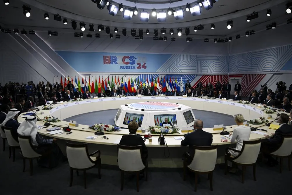 Reunião de cúpula do BRICS em 2024, realizada na Rússia, com líderes e delegados sentados ao redor de uma mesa circular. Ao fundo, bandeiras de diversos países participantes estão dispostas em sequência. A sala está bem iluminada, com uma estrutura moderna e decorada com elementos geométricos nas paredes.