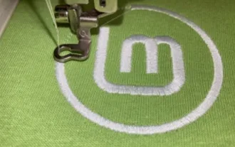 Imagem do bordado no tecido verde: "Logotipo do Linux Mint sendo bordado em tecido verde, em uma máquina de bordado."