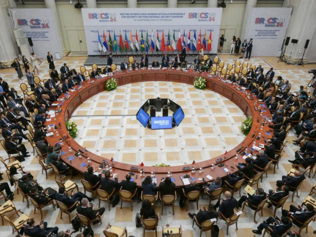 Reunião de cúpula do BRICS em 2024, realizada na Rússia, com líderes e delegados sentados ao redor de uma mesa circular. Ao fundo, bandeiras de diversos países participantes estão dispostas em sequência. A sala está bem iluminada, com uma estrutura moderna e decorada com elementos geométricos nas paredes.