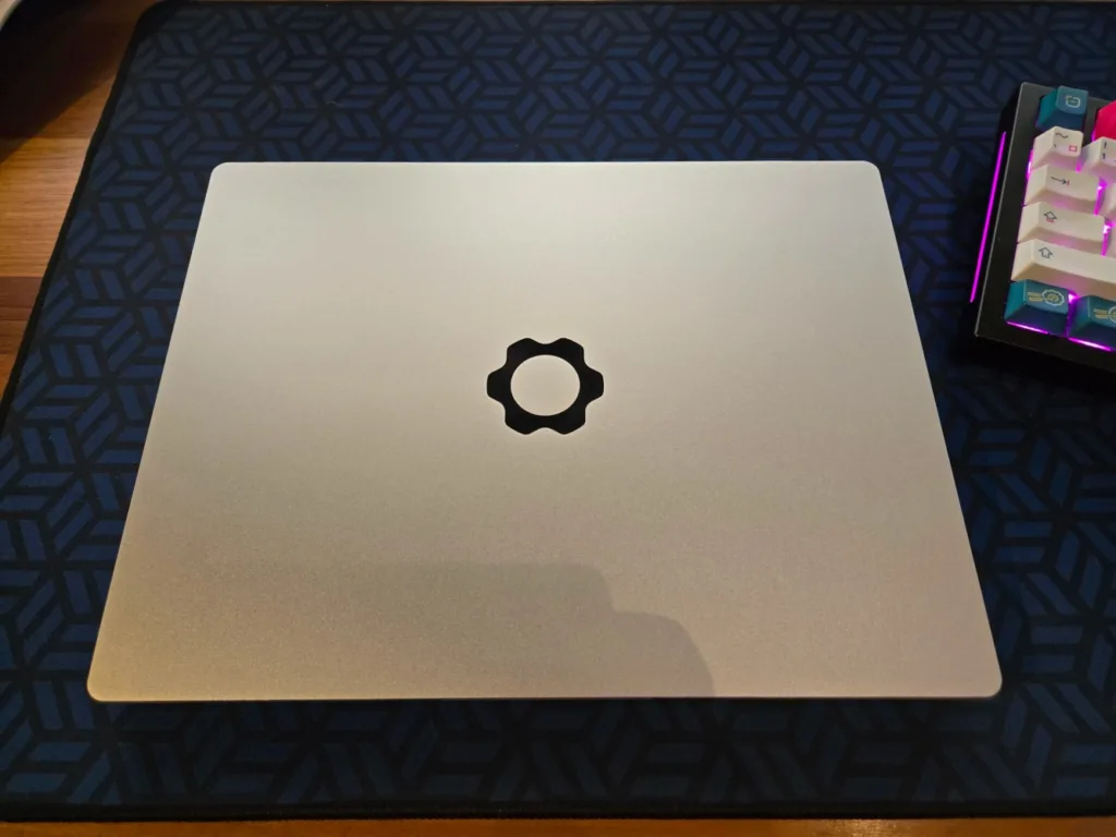 Imagem do laptop: "Laptop Framework fechado com logotipo centralizado na tampa, em uma superfície com padrão geométrico azul e preto ao fundo."