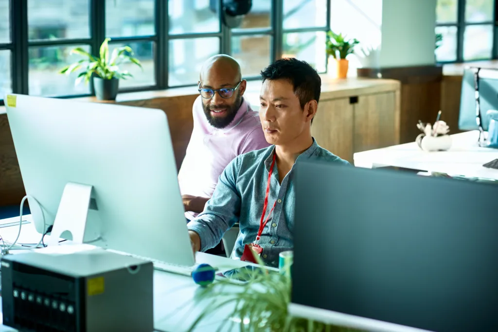 Mid adult Chinese man sitting at desk using computer, IT supporter assisting office worker, software developer looking at screen and concentrating