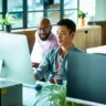 Mid adult Chinese man sitting at desk using computer, IT supporter assisting office worker, software developer looking at screen and concentrating