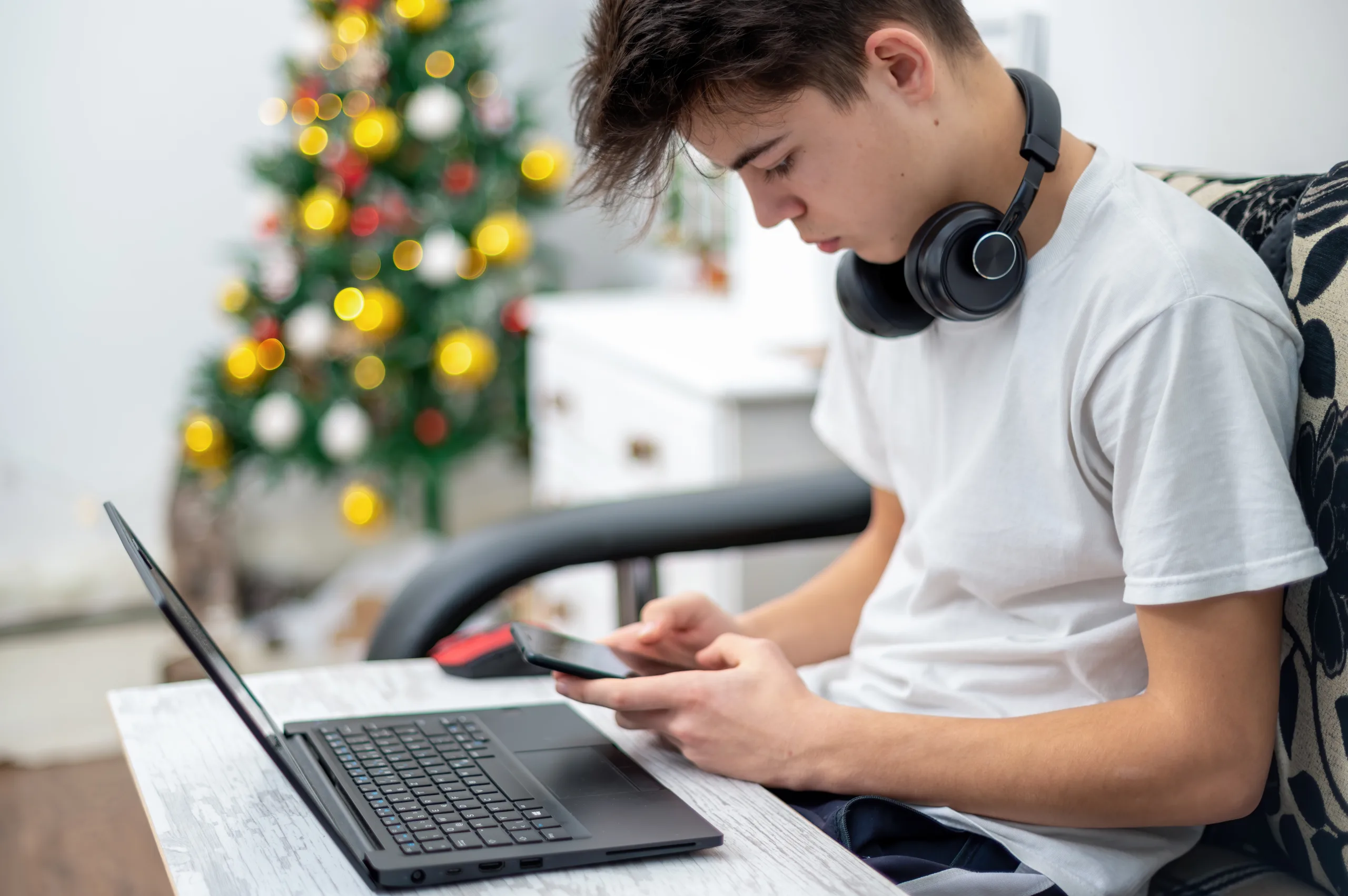 Teen boy is using smartphone with headphones, laptop on the knees at home. Christmas tree on the background. Serious and focused face
