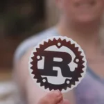 Pessoa segurando adesivo com o logotipo oficial da linguagem Rust, simbolizando o envolvimento com a programação em Rust no Linux.