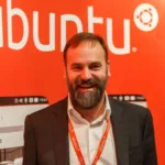 Mark Shuttleworth, pai do Ubuntu, sorrindo diante de painel com o logotipo da distribuição Linux.