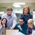 Gestor de TI orienta equipe de desenvolvedores durante code review, ilustrando colaboração e soft skills no ambiente corporativo.