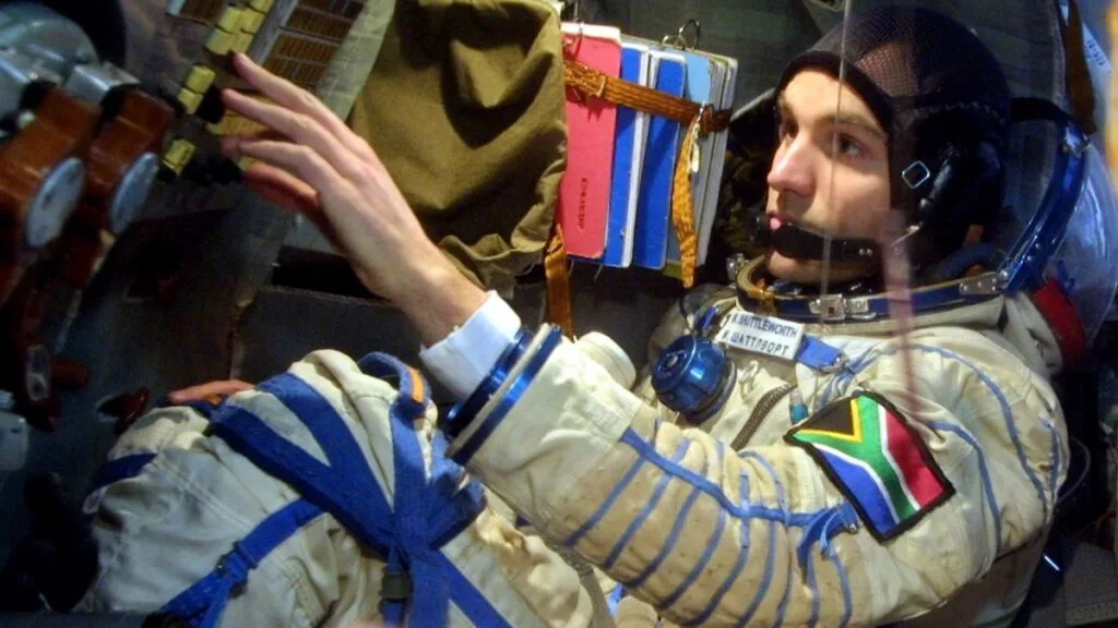 Mark Shuttleworth dentro da Soyuz, operando instrumentos com traje espacial, exibindo a bandeira da África do Sul.