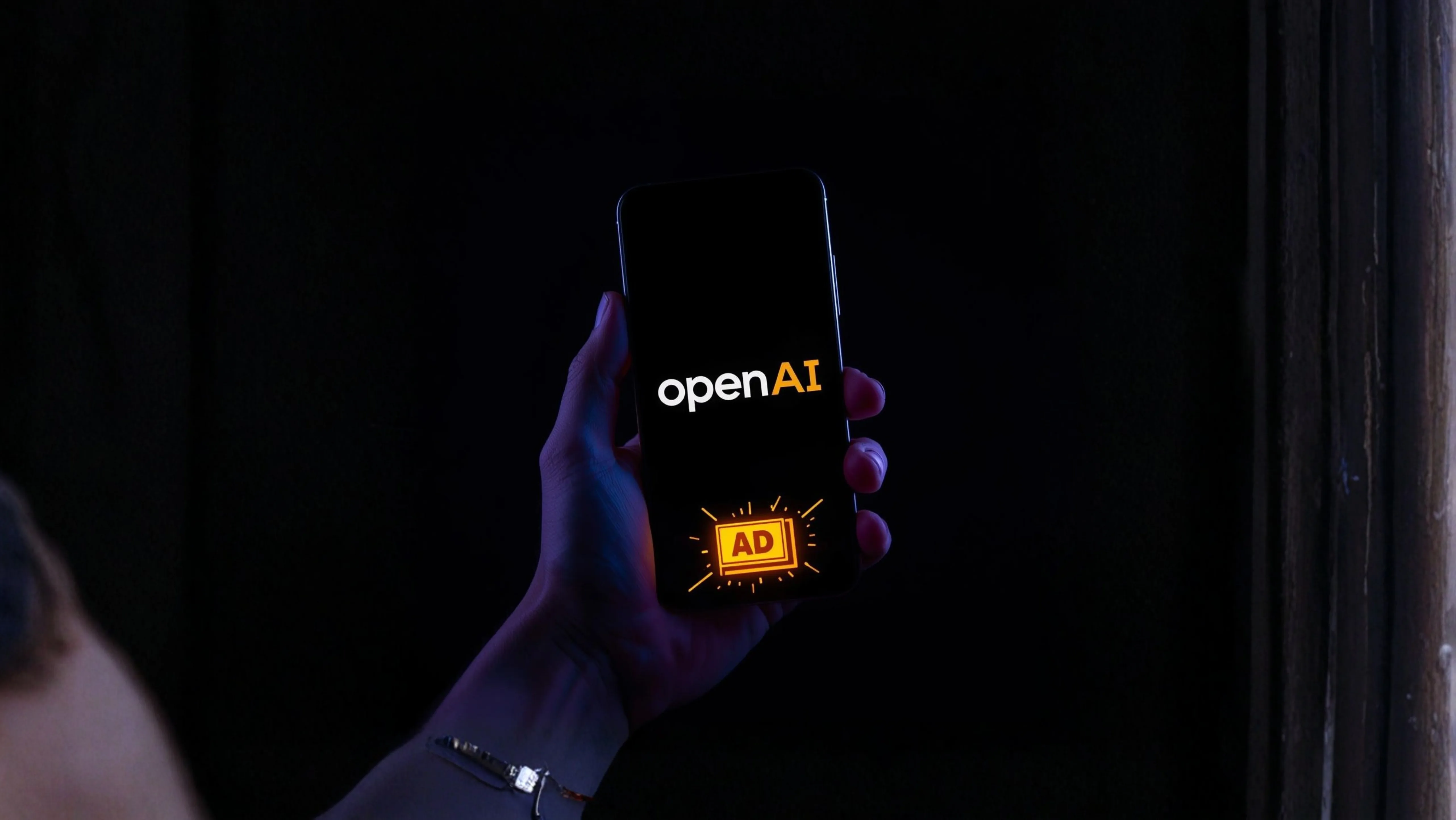 Mão segurando um smartphone em modo escuro com o logo da OpenAI e um selo “AD” em amarelo na tela